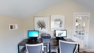Located at 2355 Michigan Ave in Pensacola, Florida, this single-floor business center provides a practical and comfortable environment designed to meet essential office needs. The space features a dedicated workstation area equipped with dual computer monitors and a multifunction printer, making it well-suited for tasks such as browsing, printing, and general office work. The room is finished with a sloped ceiling that adds architectural character and contributes to a cozy, intimate atmosphere. Neutral, light-toned walls create a clean and focused setting, while coastal-themed framed artwork depicting sea coral adds a touch of local charm and visual interest. The workstations are arranged side by side along a sturdy desk surface, each accompanied by ergonomic seating for user comfort. Natural light filters into the space through a window, keeping the environment bright and inviting. The overall design balances functionality with a relaxed, coastal aesthetic, making it a convenient and pleasant space for guests or visitors looking to handle business tasks efficiently.