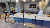 Located at 200 Henry Hill St in Cary, NC, this single-floor amenity space offers a thoughtfully designed environment that blends social gathering areas with functional fitness facilities. The social lounge immediately captures attention with its bold blue-and-white checkerboard tile flooring, soaring double-height ceilings, and expansive black-framed windows that flood the space with natural light and outdoor views. A long marble-topped bar island anchors the lounge, complemented by playful rope swings serving as seating, globe pendant lighting, and a dramatic open staircase with a green accent wall. Comfortable banquette seating lines the windowed perimeter, paired with round marble tables and warm wooden chairs, creating an inviting café-style atmosphere. An adjacent kitchenette area features dark wood cabinetry, under-counter refrigerators, a sink, and a mounted flat-screen TV set against a textured brick-tile accent wall. A cozy nook with open shelving and colorful accents adds a casual lounge zone. The fitness center is a standout feature, boasting double-height ceilings, floor-to-ceiling steel-framed windows, and a comprehensive selection of strength and cardio equipment, including weight machines, free weights, benches, plyometric boxes, and a cable system. Full-length mirrors and an upper-level overlook enhance the spacious, light-filled workout environment.