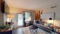 The Maple floor plan in Memphis, TN is a thoughtfully designed 2-bedroom, 2-bathroom single-level residence that blends contemporary style with everyday functionality. The living room serves as the heart of the home, featuring rich wood-look flooring, crown molding, and a striking wood slat accent wall that adds warmth and character. A sliding glass door fills the space with natural light and provides access to an outdoor area. The adjacent dining area is defined by a decorative geometric mirror wall panel and offers ample room for entertaining. The kitchen is outfitted with stainless steel appliances, white shaker cabinetry, dark granite-style countertops, and a peninsula that opens toward the dining space, creating a seamless flow. Both bedrooms are generously sized with plush carpet flooring, large windows that invite natural light, and serene, neutral color palettes. The bathrooms feature wood-look flooring, white vanity cabinetry, and glass-enclosed showers. A dedicated in-unit laundry room with a full-size washer and dryer adds convenience. Arched hallway details and consistent crown molding throughout lend an elegant, cohesive finish to this well-appointed residence.