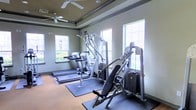 This fitness center offers a clean, functional environment designed to support a well-rounded workout routine. The space is outfitted with a selection of cardio machines, including multiple treadmills, as well as a multi-station strength training machine and a cable/weight apparatus, providing users with options for both aerobic and resistance training. Equipment is positioned on protective rubber floor mats over hardwood-style flooring, ensuring both safety and durability. The room is brightened by multiple large windows along the walls, allowing ample natural light to fill the space and offering views of the surrounding exterior. Ceiling fans are mounted above, promoting air circulation and a comfortable workout environment. The walls are finished in a muted green tone with dark crown molding accents near the ceiling, lending the space a polished, intentional aesthetic. The tray ceiling detail adds a touch of architectural interest, elevating the overall design beyond a purely utilitarian setting. The layout is organized and spacious enough to accommodate multiple users simultaneously, making it a practical and inviting amenity for daily fitness needs.