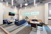 Located at 555 Boulevard NE in Atlanta, GA, this single-floor community lounge is a thoughtfully designed multipurpose space that seamlessly blends industrial aesthetics with modern comfort. Soaring ceilings with exposed ductwork and cascading linear pendant lighting create a dramatic yet inviting atmosphere throughout the open-plan layout.

The lounge area is anchored by a plush blue leather sofa and accent chairs arranged around a marble-top coffee table, all set against a striking painted brick feature wall adorned with geometric murals in muted gold, gray, and blue tones. A wall-mounted flat-screen TV and a shuffleboard table make this zone ideal for relaxed social gatherings.

A central dining area features a round marble-top pedestal table surrounded by upholstered chairs with brass-accented legs, positioned beneath a cluster of vertical pendant lights. Adjacent to this is a well-appointed kitchen area centered around a large white waterfall island with bar stool seating, complemented by a herringbone-tiled accent wall, ample counter space, and integrated appliances.

Horizontal transom windows along the walls allow natural light and greenery views to filter in, while large-format tile flooring and patterned area rugs define each zone. Glass-partitioned entry doors and lush potted plants add a polished, welcoming finish to this vibrant community space.