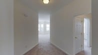 This single-floor space is defined by a clean, neutral aesthetic that creates a bright and welcoming atmosphere throughout. The interior is finished with wall-to-wall beige carpeting that runs continuously from the hallway into the adjoining living area, providing a cohesive and comfortable feel underfoot.

A hallway serves as the central connector within the space, featuring crisp white walls, white baseboards, and a flush-mount ceiling light fixture that provides overhead illumination. The hallway leads naturally into a larger living area visible at the far end, where multiple windows allow generous amounts of natural light to fill the room, enhancing the open and airy quality of the space.

The living area benefits from what appears to be a multi-window layout, flooding the interior with daylight and creating a bright, inviting environment. A door is partially visible to the right side of the hallway, suggesting access to an additional room. The overall design is straightforward and functional, with a neutral color palette of white and light beige tones throughout, making the space feel clean, versatile, and move-in ready.