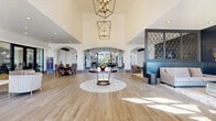 This expansive single-floor clubhouse in Plano, Texas, is a showcase of refined design and thoughtful functionality. The grand entrance foyer sets an impressive tone with soaring ceilings, floor-to-ceiling arched windows that flood the space with natural light, and a striking round accent table centered on a geometric patterned inlay—anchored beneath an elegant geometric chandelier. Wide-plank wood-look tile flooring flows seamlessly throughout, unifying each distinct zone of the space. The open living area features dramatic double-height ceilings adorned with decorative mirrored panels and ornate pendant lighting, complemented by plush seating arrangements and a mounted flat-screen display. A full-length glass wall opens views to an outdoor pool area, bathing the lounge and dining zones in abundant natural light. The kitchen and bar area is appointed with a large center island, bar seating with upholstered stools, marble-style backsplash, and sculptural pendant fixtures. A cozy lounge nook, defined by a bold dark accent wall and latticed decorative screen, offers an intimate retreat. Additional areas include a glass-enclosed conference room and casual seating alcoves, rounding out this sophisticated and versatile community space.
