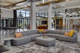 Located at 2700 W Cobalt Dr in Meridian, Idaho, this single-floor amenity center is a thoughtfully designed community gathering space that blends modern architecture with inviting, functional interiors. The space is defined by soaring double-height ceilings, exposed structural beams with warm wood accents, and polished concrete floors that flow seamlessly throughout.

Floor-to-ceiling glass walls and large sliding doors flood the interior with abundant natural light and provide expansive views of the surrounding outdoor courtyard and community grounds. Exposed brick columns add an industrial contrast to the clean, contemporary palette.

The open main hall serves as a central hub, anchored by a grand staircase and furnished with communal seating and tables suited for casual gatherings or coworking. A fully equipped demonstration kitchen features light wood cabinetry, a professional-grade stainless steel range, open shelving with tile backsplash, and a striking terrazzo island with bar seating — ideal for community events and social entertaining.

A dedicated game room includes a ping pong table, foosball table, shuffleboard, and mounted display screens for entertainment. Multiple lounge areas offer comfortable seating arrangements with bold, colorful furnishings set against bright, window-lined walls that connect seamlessly to outdoor patio spaces.