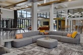 Located at 2700 W Cobalt Dr in Meridian, Idaho, this single-floor amenity center is a thoughtfully designed community gathering space that blends modern architecture with inviting, functional interiors. The space is defined by soaring double-height ceilings, exposed structural beams with warm wood accents, and polished concrete floors that flow seamlessly throughout.

Floor-to-ceiling glass walls and large sliding doors flood the interior with abundant natural light and provide expansive views of the surrounding outdoor courtyard and community grounds. Exposed brick columns add an industrial contrast to the clean, contemporary palette.

The open main hall serves as a central hub, anchored by a grand staircase and furnished with communal seating and tables suited for casual gatherings or coworking. A fully equipped demonstration kitchen features light wood cabinetry, a professional-grade stainless steel range, open shelving with tile backsplash, and a striking terrazzo island with bar seating — ideal for community events and social entertaining.

A dedicated game room includes a ping pong table, foosball table, shuffleboard, and mounted display screens for entertainment. Multiple lounge areas offer comfortable seating arrangements with bold, colorful furnishings set against bright, window-lined walls that connect seamlessly to outdoor patio spaces.