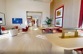 This 2,680-square-foot single-story space is a thoughtfully designed leasing lounge or community amenity center that blends contemporary architecture with warm, curated interiors. The space is anchored by a dramatic main lounge featuring soaring ceilings with expansive floor-to-ceiling windows that flood the interior with abundant natural light while framing lush outdoor views of palm trees and landscaped grounds. Light hardwood flooring flows consistently throughout, complemented by natural wood paneling, warm white walls, and bold red accent furnishings that create a cohesive, modern aesthetic. A central reception or leasing area features a sleek wood desk positioned before a panoramic window wall, offering an open and welcoming atmosphere. Adjacent seating areas include curved sofas, swivel chairs, and circular coffee tables arranged on woven area rugs, ideal for relaxed conversation. A built-in banquette with upholstered wall panels and integrated lighting provides an intimate lounge nook. Glass-walled private offices or meeting rooms with vibrant red interiors and full-height wood-framed partitions offer functional workspace. Additional areas include a conference room with an oval table and woven chairs, and a presentation or collaboration space with dual oval tables set against a slatted wood feature wall displaying curated artwork. The overall ambiance is polished, bright, and welcoming.