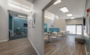 This well-appointed single-floor amenity space is designed with a cohesive modern aesthetic, blending functionality with inviting style throughout. The centerpiece is a spacious lounge and social area, featuring warm wood-look flooring, an abundance of natural light from overhead skylights, and large sliding glass doors that connect the interior to the outdoors. Comfortable seating arrangements include teal upholstered booth-style banquettes and an array of round tables with sleek white chairs, making the space ideal for both casual gatherings and focused work. Abstract teal-toned artwork adorns the crisp white walls, reinforcing the space's vibrant yet clean color scheme. A dedicated workspace nook offers counter-height desks and additional seating for productivity. The fitness center is thoughtfully equipped with cardio machines including treadmills, stationary bikes, and an elliptical, as well as a multi-station weight rack, dumbbells, and a large wall mirror. Bold teal accent walls and patterned geometric carpet in the fitness area create an energizing atmosphere, complemented by natural light from high clerestory windows. A compact, well-designed kitchen rounds out the amenities, outfitted with white cabinetry, a wall oven, an induction cooktop, a microwave, and ample storage, offering a practical space for meal preparation.