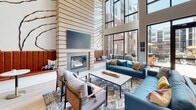 Located at 1601 Overbrook Rd in Richmond, Virginia, this contemporary two-story amenity space is designed to foster community and comfort. The main level makes an immediate impression with soaring double-height ceilings and floor-to-ceiling black-framed windows that flood the interior with natural light and provide sweeping views of the outdoor courtyard below, complete with seating and colorful umbrellas. A dramatic floor-to-ceiling fireplace clad in textured tile serves as the focal point of the main lounge, which is furnished with teal and leather sofas, patterned area rugs, and carefully curated décor that strikes a balance between modern and inviting. Adjacent to the lounge, a dedicated dining area features a large communal table with banquette seating and continued courtyard views. A well-appointed kitchenette offers marble-look quartz countertops, slate-blue cabinetry, stainless steel appliances, and bar-height seating, making it ideal for gatherings. A cozy secondary lounge area with built-in shelving and a wall-mounted display provides a quieter retreat. The upper level houses a billiards and games room, complete with a foosball table, bar seating, and local Richmond-themed artwork. Light wood flooring runs throughout both levels, unifying the space with a warm, cohesive aesthetic.