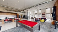 Located in Denver, CO (80249), this contemporary amenity space is designed to foster community and productivity across a single, expansive floor. The open-plan layout is anchored by soaring double-height ceilings with exposed industrial ductwork, creating a bold contrast against the crisp white walls and modern finishes throughout.

The central game lounge features a striking red billiards table beneath sculptural pendant lighting and geometric ceiling fixtures, surrounded by plush sectional seating with colorful accent chairs. Floor-to-ceiling windows flood the space with natural light and offer open views of the surrounding neighborhood and sky.

A dedicated co-working lounge offers high-backed booth seating accented in red, complemented by vibrant acoustic ceiling panels in red and gray that add both visual interest and sound management. Private work pods with glass partitions provide focused work areas nearby.

The conference room is well-appointed with a large presentation screen, warm wood cabinetry, and a sizable conference table surrounded by multi-colored rolling chairs atop a bold striped rug. A sleek kitchenette features wood cabinetry, quartz countertops, black fixtures, large-format tile flooring, and direct access to an outdoor balcony. A quiet seating nook with swivel lounge chairs rounds out the space, ideal for casual conversations or individual relaxation.