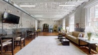 Located at 809 Chattanooga Ave in Dalton, Georgia, this single-floor space is a striking example of adaptive reuse, blending industrial heritage with thoughtful interior design. The main hall is expansive and open, anchored by soaring white-painted wood beam ceilings and original exposed brick walls that bear the character of the building's storied past — layers of paint, aged markings, and weathered textures that give the space a distinctive, authentic ambiance. Rich, wide-plank hardwood floors run throughout, adding warmth against the raw masonry backdrop. Oversized multi-pane windows flood the interior with abundant natural light and offer views of the surrounding greenery. The space is divided into distinct functional zones: a lounge area with leather seating and low coffee tables anchored by a plush area rug, and a dining and bar-height table section equipped with a wall-mounted television. A corridor with clean white walls and decorative arched brick doorways connects to additional rooms beyond. A striking open staircase with a dark wood railing serves as a central architectural focal point. The combination of industrial materials, generous proportions, and curated furnishings makes this a versatile and visually compelling space.