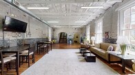 Located at 809 Chattanooga Ave in Dalton, Georgia, this single-floor space is a striking example of adaptive reuse, blending industrial heritage with thoughtful interior design. The main hall is expansive and open, anchored by soaring white-painted wood beam ceilings and original exposed brick walls that bear the character of the building's storied past — layers of paint, aged markings, and weathered textures that give the space a distinctive, authentic ambiance. Rich, wide-plank hardwood floors run throughout, adding warmth against the raw masonry backdrop. Oversized multi-pane windows flood the interior with abundant natural light and offer views of the surrounding greenery. The space is divided into distinct functional zones: a lounge area with leather seating and low coffee tables anchored by a plush area rug, and a dining and bar-height table section equipped with a wall-mounted television. A corridor with clean white walls and decorative arched brick doorways connects to additional rooms beyond. A striking open staircase with a dark wood railing serves as a central architectural focal point. The combination of industrial materials, generous proportions, and curated furnishings makes this a versatile and visually compelling space.