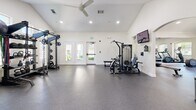 Located at 801 E Atherton Dr in Manteca, California, this fitness center offers a comprehensive workout facility spread across a single level. The space is divided into two dedicated exercise rooms, each designed to support a variety of training needs, complemented by a full bathroom for convenience.

The main exercise room is spacious and airy, featuring soaring vaulted ceilings with recessed lighting and crown molding that lend an upscale feel. A row of treadmills lines the wall beneath a series of elegant arched windows that frame lush outdoor views, filling the room with abundant natural light. Full-length mirrors visually expand the space while a wall-mounted television adds a modern touch. Durable rubber flooring runs throughout, providing safety and comfort underfoot.

The second exercise room is equipped for strength training, housing a power rack with weight plates, cable machines, and a dumbbell rack with an accompanying bench. This room also benefits from vaulted ceilings, a ceiling fan, and direct exterior access through glass-paneled doors. Spinning bikes provide an additional cardio option.

The bathroom is finished with warm wood cabinetry, a vanity with a mirror, and classic hexagonal tile flooring, rounding out this well-appointed fitness facility.