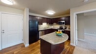 This single-floor residence at 8601 Edinbrook Crossing in Brooklyn Park, Minnesota, offers a thoughtfully designed layout with a cohesive blend of warm tones and practical finishes throughout. The kitchen is a standout feature, boasting rich dark cabinetry, granite countertops, a curved island, hardwood-style flooring, and stainless steel appliances, including a range, microwave, and dishwasher. The open sight line from the kitchen to the living area creates a connected, functional flow ideal for everyday living. The living area is generously proportioned, featuring plush carpeting, neutral walls, a textured ceiling, and a large window alongside a exterior door that invites natural light and outdoor access. The bedrooms are comfortably sized, each with soft carpet flooring, windows with blinds, and clean white trim. Two bathrooms serve the unit: one features a tub-shower combination with a stylish curtain, dark vanity cabinetry, granite countertops, and tile flooring, while the second bathroom includes a walk-in shower with glass enclosure and a matching dark vanity with granite countertop. The overall ambiance is clean, modern, and move-in ready, with consistent warm neutral tones and quality finishes that lend a welcoming feel throughout the space.