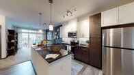 This single-floor residence showcases a thoughtfully designed open-concept layout that seamlessly connects the kitchen, dining, and living areas. The kitchen is a standout feature, boasting dark granite countertops, a central island with pendant lighting, a classic white subway tile backsplash, and a two-tone cabinetry design pairing crisp white upper cabinets with rich dark wood lower cabinets. Stainless steel appliances, including a refrigerator, electric range, and microwave, complete the space. Track lighting and stylish hanging fixtures provide ample illumination throughout.

The living area is bright and inviting, anchored by a bold navy accent wall and large sliding glass doors that open onto a private balcony, allowing generous natural light and outdoor views. The bedroom offers a serene retreat with a neutral toned accent wall, plush carpeting, and multiple windows that bring in natural light. A walk-in closet provides convenient storage.

The bathroom features dark wood vanity cabinetry with a granite countertop, a large mirror, and a full tub and shower combination with white subway tile surround and wood-look flooring. A dedicated in-unit laundry area houses a full-sized side-by-side washer and dryer with wire shelving above for added storage. Wood-look flooring flows throughout the main living spaces, tying the design together cohesively.