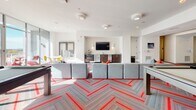 Located on 20th Street South in Birmingham, Alabama, this single-floor amenity lounge is a thoughtfully designed social space that blends bold aesthetics with practical functionality. The open-plan layout is anchored by a striking herringbone-patterned carpet with vivid red accents, creating a dynamic visual foundation throughout the space.

The lounge is divided into distinct yet connected zones. A generous seating area features large modular gray sectionals trimmed in red, paired with round wooden coffee tables and sleek white side tables, encouraging both casual conversation and relaxed lounging. A colorful horizontal striped wall panel serves as a bold focal point, adding a layer of artistic character to the environment.

A dedicated recreation zone includes a billiards table set against a geometric, artistically rendered accent wall, providing an engaging activity area. Floor-to-ceiling windows and glass doors flood the interior with natural light and offer views of an outdoor patio area, seamlessly blending indoor and outdoor living.

A built-in kitchenette is equipped with granite countertops, dark wood cabinetry, a compact refrigerator, microwave, espresso machine, and a wall-mounted flat-screen television — making it a convenient hub for residents. Café-style seating with white and yellow chairs lines the perimeter near the windows, offering additional spots for dining or socializing. Recessed and decorative ceiling lighting completes the bright, welcoming ambiance throughout.