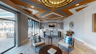Located at 3908 Independence Blvd in Wilmington, NC, this single-floor space delivers an impressive blend of coastal elegance and contemporary design. The most striking architectural feature is the elaborately crafted ceiling, which combines warm natural wood planking with geometric coffered detailing and painted gray accents. A grand circular chandelier with gold-toned cylindrical elements anchors the lounge area, while a multi-arm industrial-style pendant light illuminates the dining space below.

The open-plan layout flows effortlessly between distinct functional zones. The lounge area features wood-framed accent chairs dressed in blue shibori-patterned cushions, arranged around a live-edge coffee table atop a patterned area rug. Large sliding glass doors allow abundant natural light and provide direct access to an outdoor deck or terrace beyond.

The dining area accommodates a generous group, centered around a substantial wood-and-steel table surrounded by upholstered white chairs. Behind it, a built-in wet bar with gray subway tile backsplash, under-counter refrigeration, and a wall-mounted television adds functionality. A separate lounge nook features bold blue shiplap walls, a mounted television, and casual seating. Light wood-look flooring and neutral gray walls unify the entire space with a refined coastal ambiance.