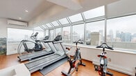 Located at 3470 Rue Stanley in the heart of Montréal, this fitness facility offers a thoughtfully designed exercise space that combines functionality with an inspiring atmosphere. The single-floor gym is characterized by warm hardwood flooring throughout and a striking glass curtain wall with skylights that flood the space with abundant natural light while framing panoramic views of the Montréal skyline.

The exercise room is comprehensively equipped to support a full range of workouts. Cardio equipment includes Matrix treadmills, an elliptical trainer, and spin bikes, all positioned to take advantage of the expansive city views. A Matrix functional trainer cable machine anchors the strength training area, complemented by a full dumbbell rack and a dedicated free weights zone with a workout bench. A medicine ball rack adds further variety for functional fitness training.

A split-level layout creates a natural zoning of the space, with a raised platform area featuring a decorative mural of the Montréal cityscape. A wall-mounted air conditioning unit ensures year-round climate comfort. The bright, airy ambiance created by the angled skylight ceiling and floor-to-ceiling windows makes this gym an energizing environment. Workout reference charts are posted on the walls, and the overall design strikes a balance between practical utility and an elevated, modern aesthetic.
