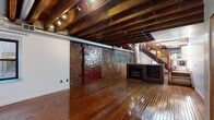 Located at 219-229 Vine St in Philadelphia's historic Old City neighborhood, this two-floor space showcases a compelling blend of industrial and historic architectural character. The lower level is defined by its raw, warehouse-inspired aesthetic: richly polished hardwood floors stretch beneath exposed wood beam ceilings fitted with track lighting, while original brick walls add texture and depth. A freestanding fireplace serves as a central focal point on this floor, complemented by a large sliding red door set into the brick wall — a distinctive industrial design element. Exposed ductwork runs along the ceiling, reinforcing the loft-like atmosphere. A full kitchen on this level features stainless steel appliances, warm wood cabinetry, terracotta tile flooring, and continued exposed beam ceilings, with natural light provided by ground-level windows. A wood staircase connects the floors, leading to the upper level where the atmosphere transitions to a quieter, more residential tone. The upper floor features the same gleaming hardwood floors, white walls, and rooms with built-in closet storage. A full bathroom with tile flooring is also present. Throughout both floors, the interplay of historic materials and open layouts creates a versatile, characterful space.