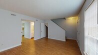 This two-story space is designed with a practical, straightforward layout that makes efficient use of every level. The main floor opens into a spacious living area finished with warm hardwood-style flooring, neutral gray walls, and a textured ceiling. Natural light enters through a large window near the front entry door, lending the area a bright and open feel. Adjacent to the living room, a dedicated dining area features matching hardwood-style flooring and is accented by a classic chandelier, with a sliding door covered by vertical blinds providing additional light. The galley-style kitchen is lined with white cabinetry on both sides, complemented by floral wallpaper accents, a double sink, electric range, dishwasher, and refrigerator — all finished in white.

A staircase with an under-stair storage closet connects the main level to the upper floor. Upstairs, the bedrooms are carpeted for comfort, each with ample natural light from windows and ceiling fans in select rooms. Closets with bi-fold doors provide convenient storage. Two bathrooms serve the space: one features a wide vanity with dual sinks, a large mirror, and a separate water closet, while the second includes a tiled tub-shower combination with dark tile flooring and a clean white surround.