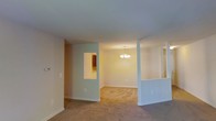 This single-floor residence offers a thoughtfully arranged open layout designed for comfortable everyday living. The main living area features wall-to-wall carpeting in neutral tones that extends throughout, creating a cohesive and warm atmosphere across the space. Soft, light-colored walls enhance the sense of openness and provide a clean, understated backdrop throughout.

The layout is defined by partial dividing walls with pass-through openings, which maintain a visual connection between the living, dining, and kitchen areas while providing a degree of separation. One pass-through reveals glimpses of wood-toned cabinetry in the kitchen, suggesting a functional and well-appointed cooking space. Another opening on the opposite side provides additional sightlines across the floor plan, contributing to the overall flow and accessibility of the interior.

The dining area is anchored by a classic multi-bulb chandelier suspended from the ceiling, adding a traditional decorative touch and providing warm ambient lighting to the space. The standard ceiling height and clean white finish maintain a straightforward, practical aesthetic throughout. White baseboards frame each room, adding a crisp finishing detail. Overall, the space presents a functional, move-in ready layout suited to a range of living needs.