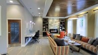 Located at 2000 Teddy House Ln in Cary, NC, this single-floor space combines functionality and contemporary design in a thoughtfully appointed interior. The open-concept layout seamlessly integrates a comfortable lounge area with a dedicated workspace, making it well-suited for both relaxation and productivity.

The lounge area serves as the focal point of the space, anchored by a sleek, modern electric fireplace set into a striking textured wall panel with an organic, wave-like design. Flanking the fireplace are built-in shelving units in a dark finish, providing display space for decorative accents. Seating throughout the lounge includes a patterned sofa and vibrant orange and yellow accent chairs, adding bold pops of color against the neutral gray walls.

A distinctive tray ceiling detail combines recessed lighting with a warm wood-paneled inset, creating visual interest overhead. Pendant lights in soft purple tones add a unique touch near the workspace, which features a long built-in desk with ergonomic seating — ideal for focused work. Natural light filters in through large windows, enhancing the welcoming, modern ambiance of the overall space.