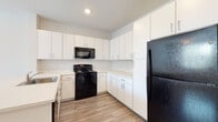 This single-floor space offers a cohesive, modern design throughout, highlighted by light wood-tone flooring that flows seamlessly from room to room. The kitchen is a standout feature, showcasing an abundance of crisp white upper and lower cabinetry with sleek chrome hardware, providing generous storage space. Light-colored countertops extend across the workspace and wrap around a peninsula with an integrated stainless steel sink, offering a practical and social layout. Black appliances — including an electric range, over-the-range microwave, and a full-size refrigerator — create a bold contrast against the white cabinetry. Recessed lighting illuminates the kitchen efficiently. The bathroom is well-appointed with a white vanity cabinet, light countertop, a large mirror, and a full tub-shower combination, all finished with the same warm wood-style flooring. The bedroom is bright and airy, featuring a window with a view of outdoor greenery, a ceiling fan with a light fixture, and neutral walls that create a calm, restful atmosphere. Throughout the unit, clean white walls, modern finishes, and consistent flooring choices contribute to a cohesive and functional living environment.