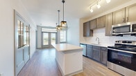 This well-designed one-bedroom, two-bathroom unit offers a thoughtfully organized single-floor layout that maximizes functionality. The open-concept kitchen and living area serves as the heart of the space, featuring light wood-tone flooring that runs throughout. The kitchen is outfitted with dark-stained shaker-style cabinetry, white subway tile backsplash, stainless steel appliances including a refrigerator, range, and built-in microwave, and a central island with a light countertop illuminated by two geometric pendant lights. A track lighting system and additional pendant fixtures provide layered illumination throughout the combined living and kitchen area.

The living area opens directly onto an outdoor balcony through a set of French doors framed by a transom window, allowing abundant natural light and outdoor views. A ceiling fan with a brushed gold finish is installed overhead. The bedroom is a comfortable size, featuring a ceiling fan, a large window with outdoor views, and access to closet space. The two bathrooms are finished with dark vanity cabinetry, white countertops, and clean white walk-in shower enclosures with brushed nickel fixtures, maintaining a consistent, contemporary aesthetic throughout the unit.