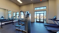 This fitness center is housed in a single-story facility with an open, airy layout accentuated by notably high ceilings and large windows that flood the space with natural light and provide views of the surrounding outdoor landscape. Double glass entry doors create a welcoming entrance, reinforcing the bright and clean aesthetic throughout.

The main workout area is generously sized and organized into distinct functional zones. A comprehensive selection of strength training equipment includes cable machines, a functional trainer, selectorized machines, and adjustable benches suited for a variety of exercises. A full dumbbell rack with multiple weight increments lines one wall, positioned in front of a large floor-to-ceiling mirror that visually expands the space and allows users to monitor their form during workouts.

The cardio zone features multiple elliptical trainers and a recumbent bike, all positioned to take advantage of the natural light provided by the large windows. The flooring throughout consists of durable rubber-style material appropriate for both heavy equipment and high-traffic use. A separate, more private room houses an additional functional trainer, offering a quieter space for focused training. A wall-mounted television and additional mirrors contribute to the overall functional and modern ambiance of this well-appointed fitness center.