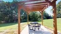 This inviting space seamlessly blends comfortable indoor living with an appealing outdoor retreat. At the heart of the outdoor area stands a well-crafted wooden pergola, constructed with warm-toned lumber and a latticed roof that filters natural sunlight. A durable round metal picnic table with attached seating sits atop a clean concrete pad beneath the pergola, framed by a backdrop of mature trees and manicured lawn — ideal for outdoor gatherings or quiet relaxation.

Inside, the living area features neutral-toned walls and carpet flooring that create a warm, understated atmosphere. A classic fireplace serves as a focal point, complemented by tasteful wall art above the mantle. Large sliding glass doors dressed with vertical blinds invite natural light and provide views of the outdoor greenery, enhancing the connection between interior and exterior spaces.

The layout is practical and well-organized, with clear sightlines from the living room toward the kitchen and adjoining hallway. The kitchen is visible with standard appliances, and closet storage is conveniently integrated near the entryway. A textured ceiling and neutral color palette throughout give the interior a clean, functional character that is both comfortable and easy to personalize.