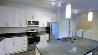 This well-appointed single-floor space showcases a contemporary open-concept kitchen designed with both style and functionality in mind. The kitchen is anchored by a full suite of stainless steel appliances, including a French-door refrigerator, an electric range, and an over-the-range microwave. Striking dark granite countertops with subtle flecks of light provide a bold contrast against the clean, white shaker-style cabinetry that extends from floor to ceiling, offering generous storage throughout. A deep stainless steel sink is set into the island or peninsula counter, paired with a modern arched faucet, making it a practical and visually appealing focal point. Overhead, a combination of recessed track lighting and an elegant pendant light provides layered illumination across the workspace. The flooring transitions between hardwood-style planks and polished concrete or tile, adding texture and warmth to the overall aesthetic. The open layout allows natural sightlines into adjacent living areas, enhancing the sense of space and connectivity. The overall ambiance is modern, bright, and clean, reflecting a thoughtfully designed interior that balances practical living with a refined, contemporary sensibility.