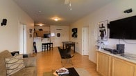 This single-floor space features an open-concept layout that seamlessly connects the living area, recreation zone, and kitchen. The main living area is anchored by light hardwood-style flooring that extends throughout, giving the space a warm, cohesive feel. A ceiling fan overhead provides ventilation and adds to the casual atmosphere of the room.

The recreation area stands out as a central feature, housing a foosball table that adds an entertaining and social element to the space. A butterfly chair and coffee table complete the casual seating arrangement, while a large gaming poster adds personality to the walls. Additional storage is provided by a wooden cabinet along one wall.

The compact kitchen is visible toward the rear and features light wood cabinetry, a wall-mounted mini-split HVAC unit, and a breakfast bar with two bar stools — ideal for casual dining or additional workspace. A front entry door is centrally positioned at the back of the main space, offering a clear sightline from the living area.

Overall, the space carries a relaxed, informal ambiance well-suited for leisure and entertainment, with functional living amenities integrated efficiently into the open floor plan.
