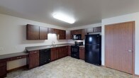 This single-floor space includes a well-appointed kitchen that serves as a central feature. The kitchen is designed with rich, dark wood cabinetry lining the walls, offering generous upper and lower storage throughout. A full suite of black appliances is included, featuring a refrigerator, electric range with overhead microwave, and a dishwasher, providing a cohesive and practical setup. The countertops are finished in a neutral tone, complementing the cabinetry, while a stainless steel sink is positioned beneath under-cabinet lighting along one wall. The L-shaped layout of the kitchen maximizes usable workspace and allows for efficient movement throughout the area. The flooring is a stone-look tile in warm, earthy tones, adding texture and durability underfoot. Overhead, flush-mount lighting provides bright, even illumination across the space. A doorway visible along the far wall suggests access to additional areas of the unit. The overall design balances functionality with a clean, modern aesthetic, making the kitchen both practical and visually cohesive for everyday use.