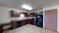 This single-floor space includes a well-appointed kitchen that serves as a central feature. The kitchen is designed with rich, dark wood cabinetry lining the walls, offering generous upper and lower storage throughout. A full suite of black appliances is included, featuring a refrigerator, electric range with overhead microwave, and a dishwasher, providing a cohesive and practical setup. The countertops are finished in a neutral tone, complementing the cabinetry, while a stainless steel sink is positioned beneath under-cabinet lighting along one wall. The L-shaped layout of the kitchen maximizes usable workspace and allows for efficient movement throughout the area. The flooring is a stone-look tile in warm, earthy tones, adding texture and durability underfoot. Overhead, flush-mount lighting provides bright, even illumination across the space. A doorway visible along the far wall suggests access to additional areas of the unit. The overall design balances functionality with a clean, modern aesthetic, making the kitchen both practical and visually cohesive for everyday use.