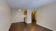 This single-floor space features an open-concept layout that connects the living area to the kitchen, creating a cohesive and functional flow throughout. The dark hardwood flooring extends across the main living space, providing a warm contrast to the neutral, light-colored walls. A textured popcorn ceiling with a ceiling-mounted light fixture adds character overhead, while white baseboards provide clean definition along the perimeter.

The kitchen is partially separated by a half-wall partition, offering a sense of division without fully closing off the space. Warm-toned wood cabinetry sits above and below the counter, and a stainless steel refrigerator is visible beyond the partition, hinting at additional kitchen space beyond. A HVAC vent is positioned high on the wall, ensuring climate control throughout the unit.

Beyond the main living area, a hallway leads to additional rooms, with shelving visible in what appears to be a storage or utility area. The overall ambiance is modest and functional, with a neutral palette that provides a versatile backdrop for a variety of interior styles.