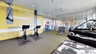 This fitness center is designed to support a comprehensive workout experience within a bright, open layout. The space is organized into distinct functional zones, seamlessly connected by a cohesive interior design. Carpet tile flooring spans the main exercise areas, providing cushioning underfoot, while recessed lighting and ceiling fans maintain a comfortable environment throughout.

The cardio zone features treadmills, ellipticals, stair climbers, and stationary cycling bikes, positioned near a large window that allows natural light to fill the room. Wall-mounted televisions offer entertainment during workouts. The strength training area includes a multi-functional cable machine system, a full rack of dumbbells with a dedicated flat bench, and various resistance attachments, ensuring options for users at every fitness level. Exercise balls are neatly stored along one wall.

A bold accent wall, painted yellow with motivational fitness-themed typography, adds visual energy to the space. Floor-to-ceiling glass panel partitions with frosted branding separate sections of the fitness center while maintaining an open, airy feel and offering views into adjacent areas. The overall ambiance is modern, energizing, and purposefully designed to encourage an active lifestyle.
