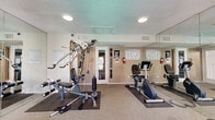 This fitness center is a dedicated, single-room exercise facility designed to accommodate a comprehensive workout experience within a compact footprint. The room is finished with neutral-toned walls, textured ceiling with recessed lighting, and low-pile carpet flooring accented by rubber equipment mats for safety and stability.

A full-length wall mirror spans one side of the room, effectively doubling the perceived size of the space and allowing users to monitor their form during exercise. The cardio section is equipped with treadmills, an elliptical trainer, and a recumbent stationary bike, all positioned along the windowed wall. Multiple windows fitted with horizontal blinds line this wall, allowing natural light to filter in while maintaining privacy, and a glass-paneled entry door provides an additional connection to the outdoors.

On the opposite side of the room, a multi-station cable weight machine offers a wide range of strength training options, complemented by free weights for added versatility. Workout reference charts and fitness center rules are posted on the walls, along with a fire extinguisher for safety compliance.

The layout is efficient and thoughtfully organized, ensuring users can move between equipment comfortably. The mirrored wall, natural light, and well-maintained equipment combine to create a functional and motivating environment suited for daily fitness routines.