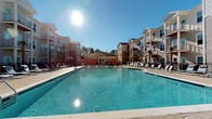 Located at 4830 Wescott Blvd in Summerville, South Carolina, this outdoor amenity space serves as a central gathering hub for the surrounding residential community. The centerpiece of the area is a generously sized resort-style swimming pool with crystal-clear blue water, designed to accommodate both relaxed leisure and active recreation. The pool deck is lined with rows of lounge chairs and chaise seating, offering ample space for sunbathing and socializing under the open South Carolina sky. A charming clubhouse or pool house structure with a warm, coral-toned exterior and French doors anchors the far end of the pool area, providing a welcoming focal point and functional support space. Palm trees and mature landscaping add a tropical ambiance throughout, complementing the sunny, warm climate of the Lowcountry region. The surrounding multi-story residential buildings feature balconies that overlook the pool area, creating a cohesive and vibrant community atmosphere. Abundant natural light floods the space throughout the day, enhancing the bright and inviting character of the environment. Overall, this amenity area reflects a well-designed outdoor living experience tailored for comfort, community, and year-round enjoyment.