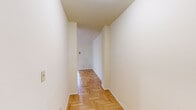 This 2-bedroom, 1-bathroom residence is thoughtfully organized on a single floor, offering a practical and comfortable layout. Distinctive herringbone-patterned hardwood floors run throughout the living areas, bedrooms, and hallways, lending a timeless character to the space.

The generously sized primary bedroom features ceiling fan lighting and two windows that invite natural light and views of surrounding greenery, creating a bright and airy atmosphere. The second bedroom is equally well-appointed, also showcasing the herringbone hardwood floors and a ceiling fan.

The kitchen is efficiently designed with white cabinetry extending to the ceiling for ample storage, white appliances including a gas range, a double sink, and a window overlooking outdoor greenery, providing natural light while cooking.

The bathroom retains a classic aesthetic with warm beige tile work, a full bathtub with shower, a pedestal sink, a decorative oval mirror, and a patterned mosaic tile floor, all illuminated by a vintage-style light fixture and a small window.

Three hallways connect the rooms seamlessly, contributing to the functional flow of the floor plan. The overall ambiance is clean, bright, and classic, with neutral white walls complementing the warm tones of the hardwood floors throughout.