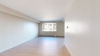 This single-floor residence offers a clean, functional layout designed for comfortable urban living. The living areas feature light hardwood-style flooring that runs throughout, complemented by neutral warm-toned walls and white ceilings that create a bright and open atmosphere. Large sliding windows in the main living spaces allow natural light to pour in while framing views of the surrounding cityscape and mature trees, adding a dynamic outdoor connection to the interior.

The galley-style kitchen is well-equipped with warm wood cabinetry, dark granite countertops, a stone tile backsplash, and a full suite of stainless steel appliances including a range, microwave, and refrigerator. Track lighting overhead provides focused illumination throughout the cooking space.

The bedrooms are finished with plush carpeting for added comfort and warmth. Each room benefits from generous window placement, with one bedroom offering a notable panoramic view of the urban skyline and autumn foliage. Mirrored sliding closet doors add a sense of depth and provide practical storage solutions.

Baseboard heating runs along the perimeter of multiple rooms, ensuring consistent warmth throughout. The overall design is understated and neutral, offering a versatile canvas well-suited to a variety of interior styles.