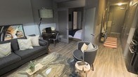 This single-floor residence showcases a carefully curated modern aesthetic defined by a rich, moody palette of deep grays and charcoal tones throughout. The open-concept layout connects the living area, workspace, and bedroom with a natural sense of flow, making efficient use of every square foot.

The living area serves as the heart of the space, anchored by a sleek glass-top coffee table resting on a dramatic marbled area rug in earthy tones. Seating includes a dark gray sofa and a stylish curved accent chair with white piping detail, adding a refined contrast to the darker surroundings. A statement floor lamp with a dark drum shade provides both ambient and task lighting.

A dedicated workspace is neatly integrated into the layout, featuring a compact dark wood desk paired with a modern chair — an ideal setup for productivity without compromising the overall design.

Wide-plank light wood flooring runs throughout, offering a warm counterbalance to the cool gray walls and dark furnishings. The bedroom, visible through a sliding or hinged interior door, continues the refined color scheme with crisp white bedding providing a clean focal point.

Overall, the space exudes a sophisticated urban character, with thoughtful furniture placement and a consistent design language that creates a polished, functional, and inviting environment.
