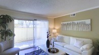 This single-floor space showcases a well-appointed living area designed for comfort and functionality. The living room is anchored by a large sliding glass door fitted with vertical blinds, which floods the space with natural light and provides direct access to an outdoor patio area. Views of mature trees and a privacy fence are visible through the glass, creating a pleasant connection to the outdoors.

The walls are finished in warm, neutral tones that complement the textured popcorn ceiling, lending the room a cozy atmosphere. Crown molding trims the upper edges of the walls, adding a refined architectural detail. A centrally positioned HVAC vent is integrated into the ceiling, ensuring year-round climate comfort.

The living space is styled with a white sofa adorned with accent pillows, paired with a sleek rectangular coffee table topped with decorative accessories. A statement vase with dried botanicals adds visual interest, while a framed botanical artwork anchors the main wall. A table lamp provides warm ambient lighting. The flooring features a plush area rug, contributing to the overall comfortable and inviting ambiance of the room.