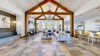 The Battlecreek Dr Clubhouse is a single-floor gathering space that masterfully blends rustic and contemporary design elements. Upon entry, visitors are greeted by a dramatic foyer with dark textured walls, wood timber framing, and large glass-paneled doors that flood the space with natural light.

The expansive main living area commands attention with its soaring cathedral ceiling, defined by bold exposed wood timber trusses and a statement chandelier. A wall of tall, multi-pane windows draws in abundant daylight and outdoor views, creating an airy, welcoming atmosphere. The multicolored slate tile flooring runs seamlessly throughout, grounding the space with an earthy, organic character.

A whitewashed brick fireplace wall serves as a striking focal point, flanked by built-in bench niches with pendant lighting — ideal for intimate seating. The generous living area accommodates large sectional sofas arranged around a modern coffee table, offering ample space for relaxed gatherings.

Framed by substantial wooden columns, the kitchen and dining area transitions naturally from the living room. The kitchen features gray shaker cabinetry, dark countertops, stainless steel appliances, and open shelving. A live-edge wood dining table paired with modern white chairs sits beneath oversized metallic pendant lights, completing a sophisticated yet approachable communal space.