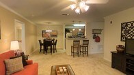 This single-floor residence offers a well-designed open-concept layout that integrates the living, dining, and kitchen areas into one cohesive and functional space. The living area is anchored by a ceiling fan with integrated lighting, providing both comfort and illumination, while neutral-toned carpeting runs throughout, creating a warm and inviting atmosphere.

The dining area is defined by an elegant chandelier suspended above a dark wood dining table surrounded by matching chairs, complemented by decorative wall art that adds a touch of personality to the space. Adjacent to the dining room, a convenient breakfast bar with seating for two bar-height chairs creates an additional casual dining option and serves as a natural transition point into the kitchen.

The kitchen is partially open to the main living area via a pass-through counter, offering glimpses of wood-toned cabinetry, stainless steel appliances, and pendant lighting that enhances the culinary space. Crown molding along the ceilings adds a refined architectural detail that carries throughout the main living areas, lending the space a polished, finished look. The overall color palette of soft beiges and warm neutrals contributes to a comfortable and cohesive ambiance ideal for everyday living.