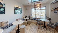 This thoughtfully designed single-floor space combines a fully equipped kitchen with a welcoming lounge and dining area. The kitchen is a standout feature, showcasing bold navy blue cabinetry accented with gold hardware, complemented by light stone countertops and a glossy subway tile backsplash. Stainless steel appliances include a French door refrigerator, wall oven, microwave, dishwasher, and a gas cooktop set into a large central island. Open shelving with white cabinetry provides additional display and storage space. The flooring throughout is a herringbone-patterned porcelain tile that lends warmth and visual interest to the entire space. The adjacent lounge and dining area is bright and airy, benefiting from large windows that invite abundant natural light and outdoor views. Round marble-top tables are paired with upholstered stools and woven chairs, while a built-in banquette seat runs along the window wall, dressed in patterned blue fabric and decorative pillows. An iron-frame candle-style chandelier anchors the seating area, and a large agate-inspired canvas artwork adds a sophisticated, contemporary touch to the overall ambiance.