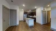 This single-floor space showcases a contemporary open-concept layout centered around a well-appointed kitchen. The kitchen is a standout feature, equipped with stainless steel appliances including a full-size refrigerator, microwave, and range. Dark espresso-toned cabinetry lines the walls, providing ample storage, while a central island with a light-toned stone countertop serves as both a prep area and a casual gathering space. The countertops throughout the kitchen complement the clean, modern aesthetic.

The flooring throughout the space consists of rich hardwood-style planks in a warm brown tone, adding warmth and visual continuity across the open layout. White walls and recessed ceiling lighting create a bright, well-illuminated environment that enhances the sense of openness. The ceiling height contributes to an airy feel throughout.

Two interior doors are visible, suggesting additional rooms beyond the main open area. The overall design is clean and contemporary, with a neutral color palette of whites, grays, and dark wood tones that provides a versatile backdrop suited to a variety of furnishing styles.