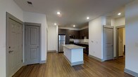This single-floor space showcases a contemporary open-concept layout centered around a well-appointed kitchen. The kitchen is a standout feature, equipped with stainless steel appliances including a full-size refrigerator, microwave, and range. Dark espresso-toned cabinetry lines the walls, providing ample storage, while a central island with a light-toned stone countertop serves as both a prep area and a casual gathering space. The countertops throughout the kitchen complement the clean, modern aesthetic.

The flooring throughout the space consists of rich hardwood-style planks in a warm brown tone, adding warmth and visual continuity across the open layout. White walls and recessed ceiling lighting create a bright, well-illuminated environment that enhances the sense of openness. The ceiling height contributes to an airy feel throughout.

Two interior doors are visible, suggesting additional rooms beyond the main open area. The overall design is clean and contemporary, with a neutral color palette of whites, grays, and dark wood tones that provides a versatile backdrop suited to a variety of furnishing styles.