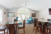 Located at 730 Franklin Rd SE in Marietta, Georgia, this single-floor space offers a thoughtfully designed open layout that seamlessly connects multiple functional areas. The standout architectural feature is a dramatic arched window that anchors the sunroom area, flooding the interior with natural light and offering pleasant views of the surrounding greenery. A ceiling fan adds comfort to this bright gathering space, which is furnished with teal wingback chairs that lend a pop of color against the neutral palette.

The dining area accommodates a round glass-top pub-height table with dark wood and leather chairs, positioned near a set of French doors that open to an outdoor patio, further enhancing the connection between interior and exterior spaces. Vaulted ceilings in this section of the space contribute to an airy, open ambiance.

The living area features a comfortable beige sofa accented with teal and patterned throw pillows, complemented by a wall-mounted flat-screen television and coordinating teal cabinetry. A dedicated workspace with a desk, computer, and teal accent lamp rounds out the functional offerings of the space. Neutral carpet flooring, soft gray walls, and a textured ceiling run throughout, creating a cohesive and welcoming environment throughout the entire floor.