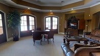 This single-floor space exudes a refined, traditional character highlighted by thoughtful architectural details throughout. The main lounge area draws the eye upward to a beautifully crafted tray ceiling adorned with decorative dentil molding, adding a sense of depth and elegance to the room. Warm, earthy tones on the walls complement the rich wood tones of the furnishings, creating a cohesive and welcoming atmosphere.

Natural light pours generously into the space through a pair of arched French doors framed with classic white trim, offering views of the surrounding exterior and brightening the neutral carpeted floors. The seating arrangement centers around a traditional dark wood table surrounded by upholstered accent chairs, ideal for casual gatherings or conversation. A leather sofa anchors a secondary seating area nearby, adding comfort and versatility to the layout.

An ornate dark wood entertainment armoire houses a flat-screen television, serving as a functional yet decorative focal point along one wall, complemented by matching furniture pieces and decorative accessories. Recessed lighting throughout ensures the space remains well-lit regardless of the time of day. Overall, this space blends classic design sensibilities with practical amenities, offering a comfortable and stylish environment for relaxation and socializing.