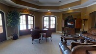 This single-floor space exudes a refined, traditional character highlighted by thoughtful architectural details throughout. The main lounge area draws the eye upward to a beautifully crafted tray ceiling adorned with decorative dentil molding, adding a sense of depth and elegance to the room. Warm, earthy tones on the walls complement the rich wood tones of the furnishings, creating a cohesive and welcoming atmosphere.

Natural light pours generously into the space through a pair of arched French doors framed with classic white trim, offering views of the surrounding exterior and brightening the neutral carpeted floors. The seating arrangement centers around a traditional dark wood table surrounded by upholstered accent chairs, ideal for casual gatherings or conversation. A leather sofa anchors a secondary seating area nearby, adding comfort and versatility to the layout.

An ornate dark wood entertainment armoire houses a flat-screen television, serving as a functional yet decorative focal point along one wall, complemented by matching furniture pieces and decorative accessories. Recessed lighting throughout ensures the space remains well-lit regardless of the time of day. Overall, this space blends classic design sensibilities with practical amenities, offering a comfortable and stylish environment for relaxation and socializing.
