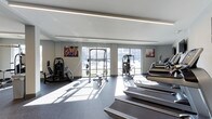 The fitness center at 333 Central Avenue is a thoughtfully designed, single-floor exercise facility that combines functionality with a clean, modern aesthetic. The space is bathed in abundant natural light streaming through large floor-to-ceiling windows along the far wall, which also provide views of the surrounding outdoor environment, creating an open and energizing atmosphere.

The interior features a neutral color palette of light gray walls and a white ceiling, complemented by sleek linear LED lighting that runs the length of the room, ensuring consistent illumination throughout. Dark rubber flooring provides both durability and safety underfoot.

The facility is comprehensively equipped to support a full range of workout routines. A power rack with a weight bench serves as a central strength-training station, accompanied by a multi-functional cable machine for resistance exercises. On the left side of the room, a selection of selectorized weight machines and a recumbent bike cater to both strength and low-impact cardio training. Along the right wall, a row of treadmills offers ample cardio capacity. Motivational fitness imagery adorns the walls, contributing to an energetic and welcoming environment.